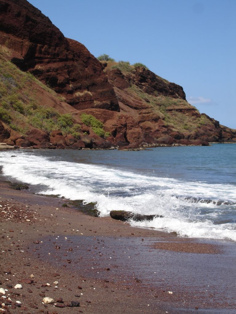 Foamy waves rolling onto dark volcanic sand beneath rugged red cliffs at Oneuli Beach.