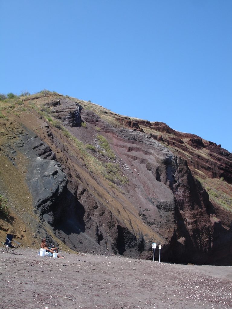 Steep layers of red, brown, and black volcanic rock rising behind Oneuli Black Sand Beach.